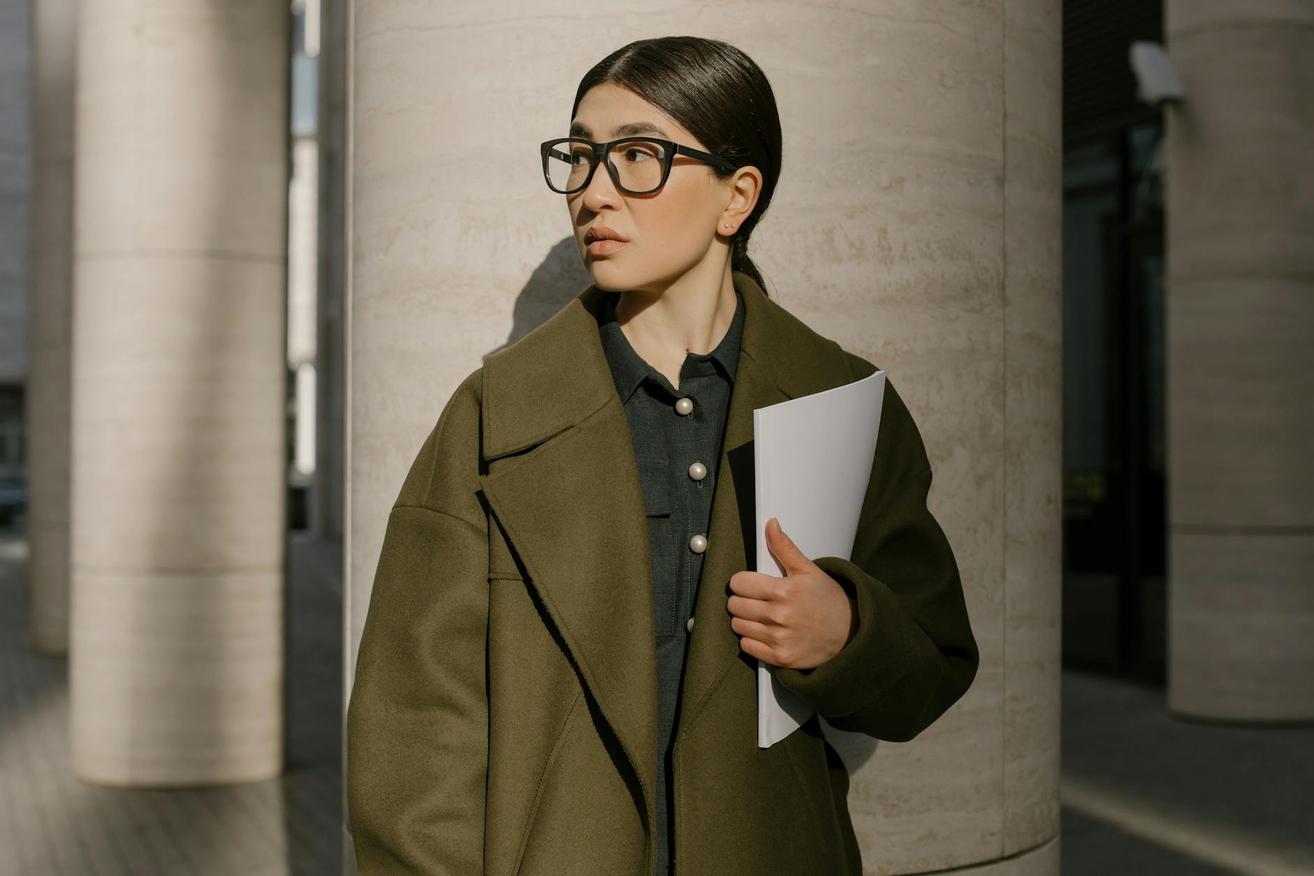 A professional woman in a coat standing outside, holding papers and looking away thoughtfully.